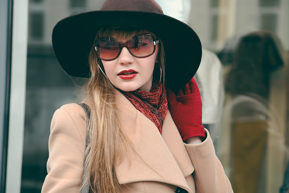 Sophisticated Winter Style – Magnolia Eye Group A fashionable woman in sunglasses, a wide-brim hat, and a beige coat poses confidently on a city street – Magnolia Eye Group.