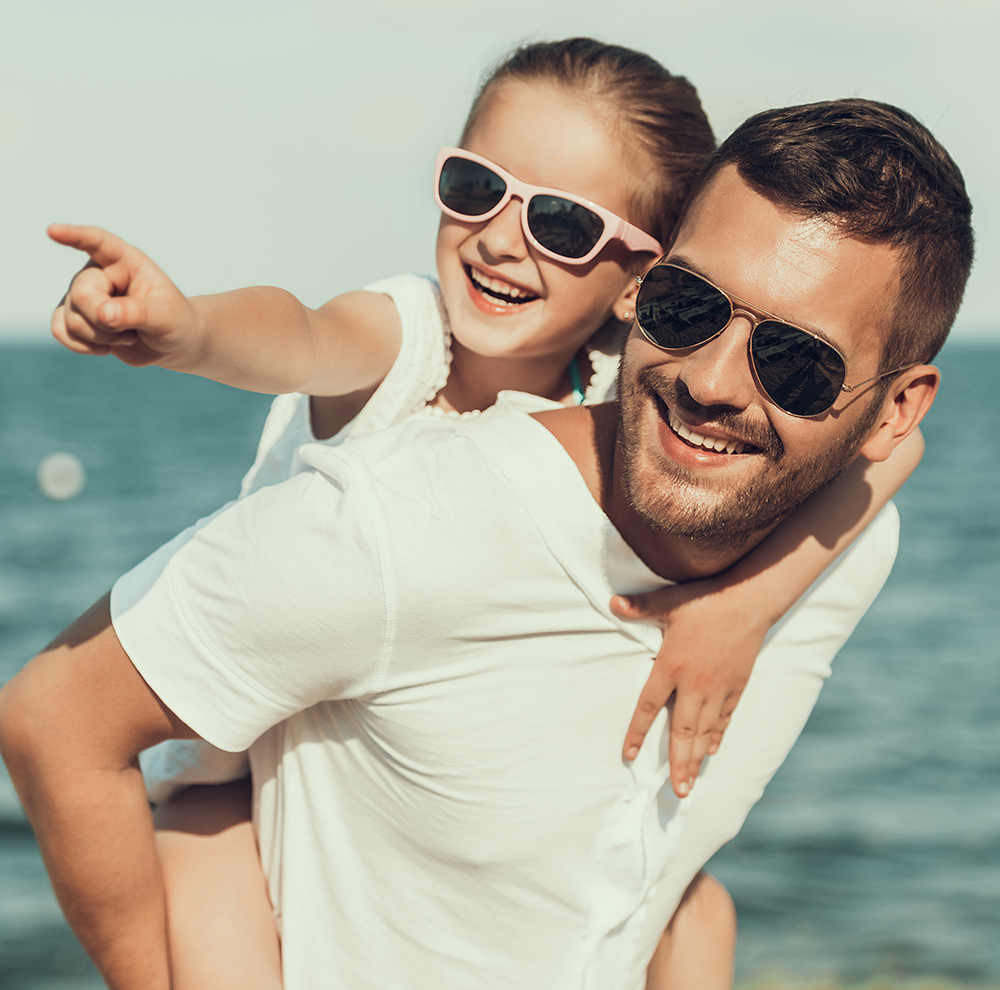 Fun in the Sun with Stylish Shades – Magnolia Eye Group A smiling man gives a piggyback ride to a young girl as they both wear sunglasses and enjoy a sunny day by the water – Magnolia Eye Group.