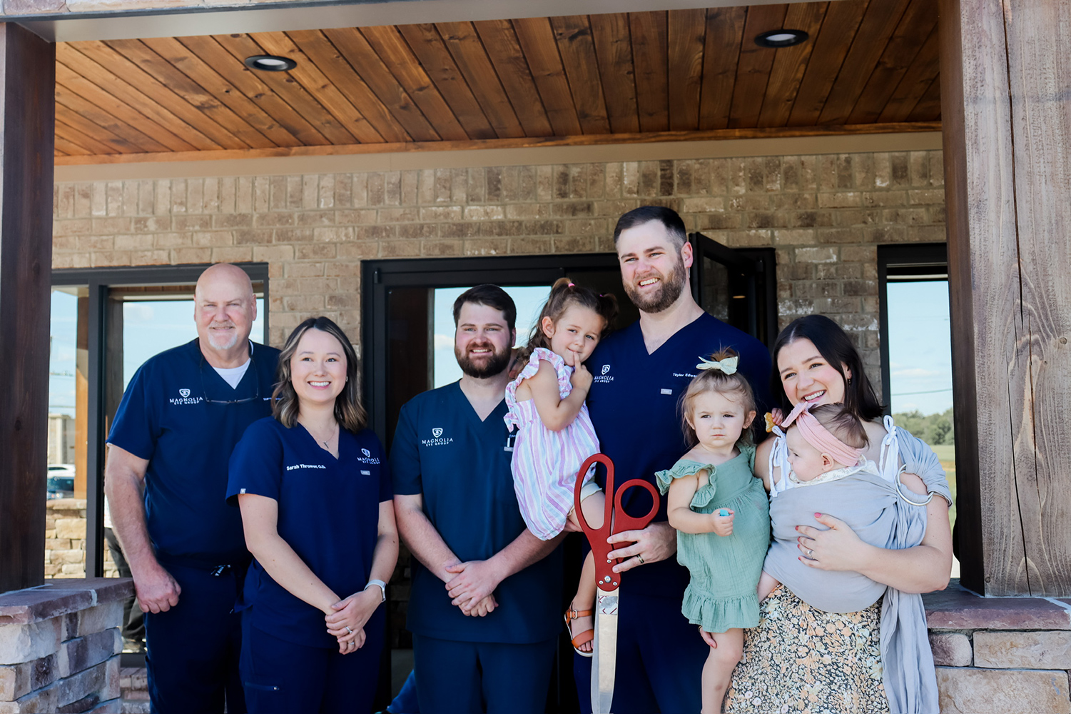 Meet the Team at Magnolia Eye Group – Magnolia Eye Group The Magnolia Eye Group team stands outside their clinic, smiling and posing with children and giant red scissors at a ribbon-cutting event - Magnolia Eye Care